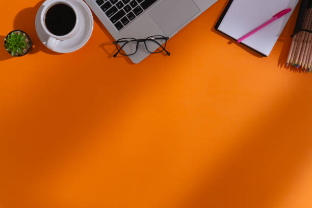 istock Home office workspace with a cup of coffee, a laptop, a pen, a notebook, an artificial plant, a pair of glasses, a bundle of colored pencils on pencil holder and shadows on orange background 1610619306