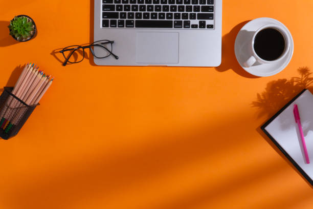 istock Home office workspace with a cup of coffee, a laptop, a pen, a notebook, an artificial plant, a pair of glasses, a bundle of colored pencils on pencil holder and shadows on orange background 1610619310