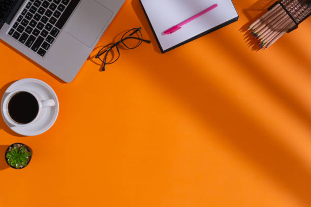istock Home office workspace with a cup of coffee, a laptop, a pen, a notebook, an artificial plant, a pair of glasses, a bundle of colored pencils on pencil holder and shadows on orange background 1610619312