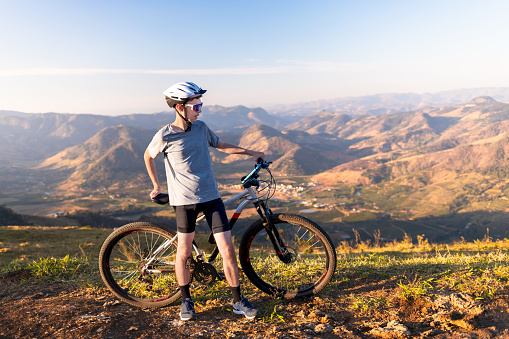 Cyclist stopped to rest beside his mountain bike,  wearing sunglasses and a helmet, with a mountain landscape in the background