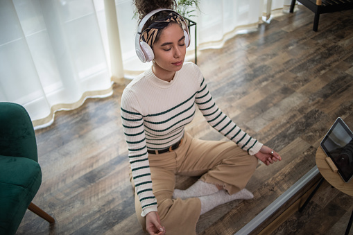 A young woman listens to a guided meditation on headphones, she is in a comfortable blue apartment