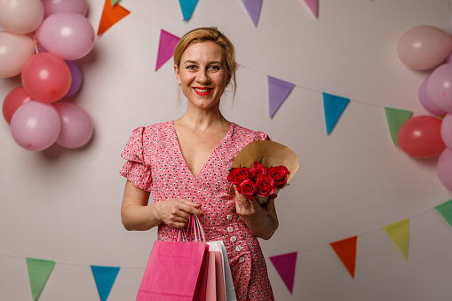 Copy space shot of charming mid adult woman standing against a white background with colorful bunting garland on the wall, holding a bouquet of red roses and gift bags that she got for her birthday. She is looking at camera and smiling happily.
