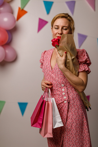 Front view of joyful mid adult woman standing against a white background with colorful bunting garland on the wall, smelling a bouquet of red roses and holding gift bags that she got for her birthday.