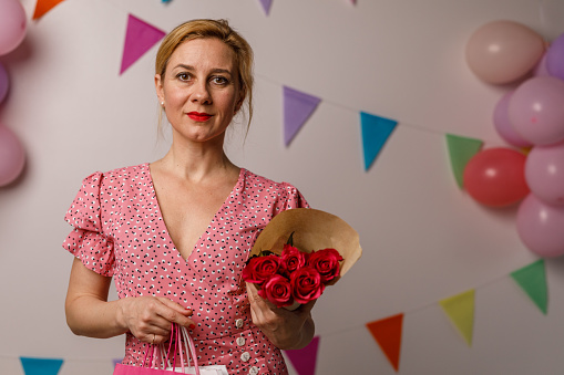 Copy space shot of charming mid adult woman standing against a white background with colorful bunting garland on the wall, holding a bouquet of red roses and gift bags that she got for her birthday. She is looking at camera and smiling happily.