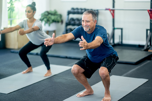 A senior gentleman stretches with his daughter in a small studio as they work to stay fit and healthy.  Both are wearing fitness attire and are focused on holding their air squat.