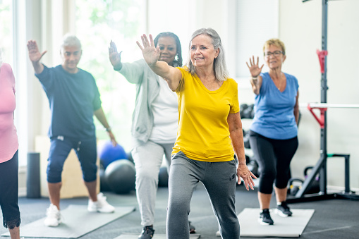 A small group of diverse seniors participate in a Tai Chi class together in a fitness studio.  They are each dressed comfortably as they work through the movements and focus on their breathing.