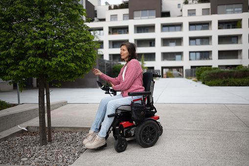 A disabled woman with a severe genetic defect (dystrophy) in a wheelchair is on the phone and looking at the green trees in the neighborhood of her apartment block.