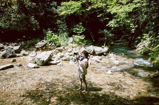 A woman walking along a river in a forest while taking pictures with a camera
