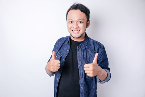 Excited Asian man gives thumbs up hand gesture of approval, isolated by white background