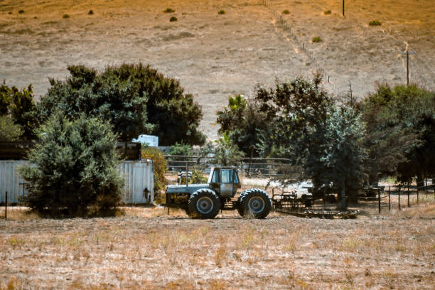 An old rusty farming tractor sits in a field of dead grass outside a white fence surrounding a farmhouse in central California.