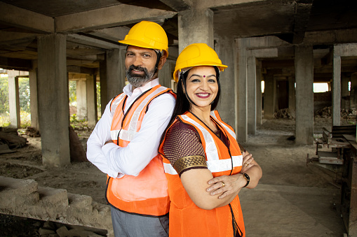 Portrait of two Indian male and female civil engineers or architect wearing helmet standing with cross arms. Real estate.
