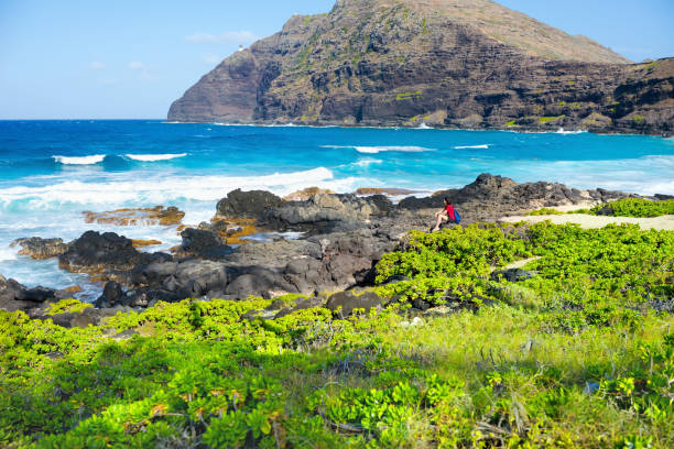 하와이 오아후섬의 마카푸우 해변에서 바위가 많은 해안선을 따라 앉아 있는 한 여성 스톡 사진