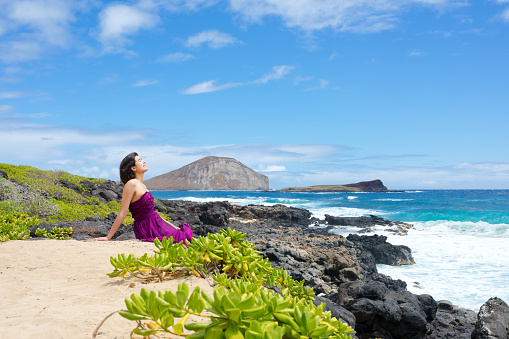 One young woman in purple dress sitting on rocky shore looking out over ocean at Makapu'u beach in Oahu, Hawaii