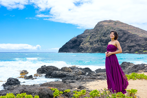 Teen girl in purple dress standing by rocky beach  on Hawaiian coast at Makapu'u beach