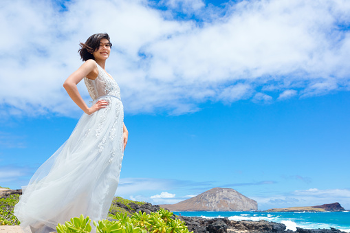 Smiling teen girl in white dress standing by rocky lava rocks on Hawaiian beach by ocean