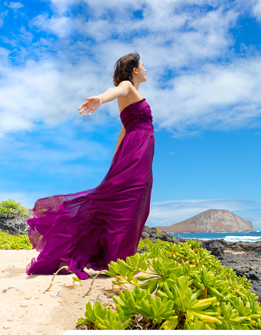 Teen girl in purple dress enjoying breeze on Hawaiian coast, arms outstretched