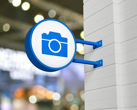 Camera icon on hanging blue rounded signboard over blur light and shadow of shopping mall, Business camera service concept, 3D rendering