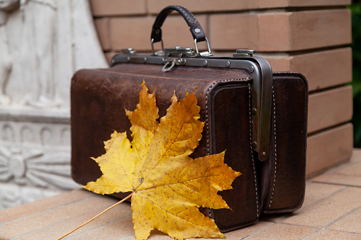 Autumn in vintage style. Vintage leather bag stands on a brick fence. On top lies a large yellow leaf of maple.