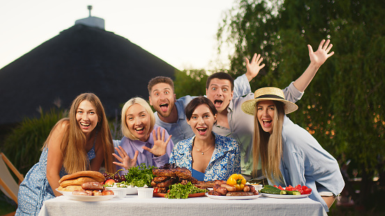 Outdoor dining table with delicious BBQ meats, fresh vegetables and salads. Happy people are dancing and having fun in the background.