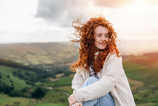 Happy smiling red hair Woman in jacket reaching the destination and sitting on the top of mountain at sunset. Travel Lifestyle concept The national park Peak District in England