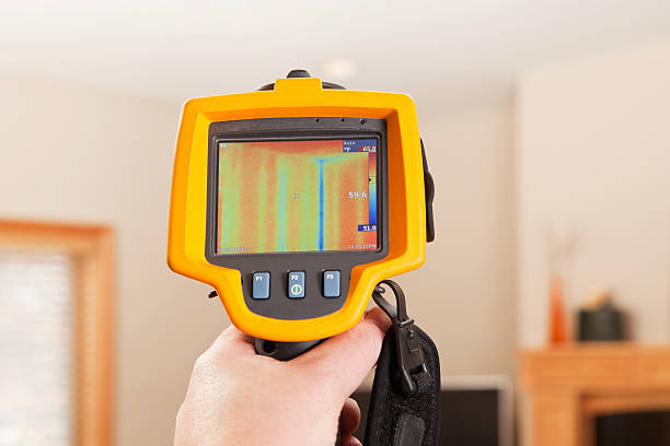 Infrared Thermal Imaging Camera Pointing to House Wall An infrared thermal imaging system being used during a home energy audit. The camera is pointed to a house living room wall and shows a distinct blue (cold) area within the home’s insulation. The center target area reads 59.6 degrees with a range of 51 to 65 degrees in the area. The blue area is a stud behind the wall, this is a typical reading in any stick built home. The wood's R-value is less than the insulation so the camera reads cooler in this area. Energy audits are performed to determine how efficient the house is and to suggest steps to increase energy efficiency. To the left is a window frame, the camera is pointed above a TV with a fireplace mantle on the right. winterizing stock pictures, royalty-free photos & images