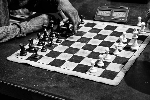 Close-up low-angle view of serious gray-haired mature male performing move with pawn piece on wooden chessboard, looking at camera. Handsome bearded man playing chess alone at home.