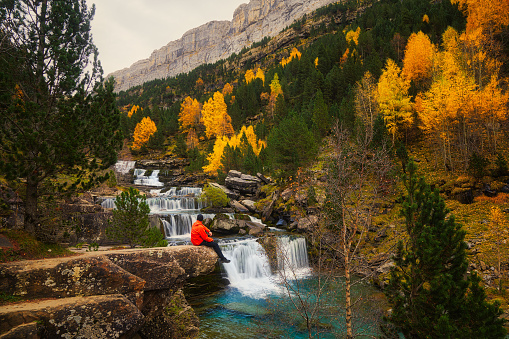 Man with an orange jacket sat on a rock in a forest in autumn looking back at waterfalls flowing