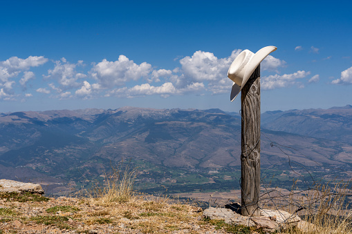 A white cowboy hat standing on a wooden pole. There's a panoramic view of mountains and white clouds in the sky