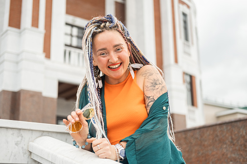 A young happy independent hipster woman with long colored dreadlocks, a nose piercing and a tattoo on her shoulder walks along a city street, looks at the camera and laughs