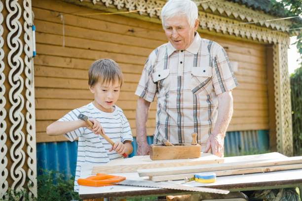 istock Elderly gray-haired mature man and a small boy are standing at a table with carpentry tools. Grandfather teaches his grandson to carpenter outdoors in the backyard of the house on a sunny day. 1611517065