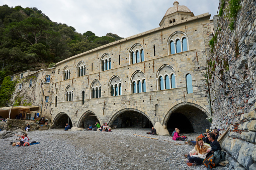 Camogli, Italy - April 03, 2022: tourists on the rocky beach in front of Abbey of San Fruttuoso, on the italian riviera between Camogli and Portofino. The abbey can only be reached by sea or by hiking trails. Genova. Italy.