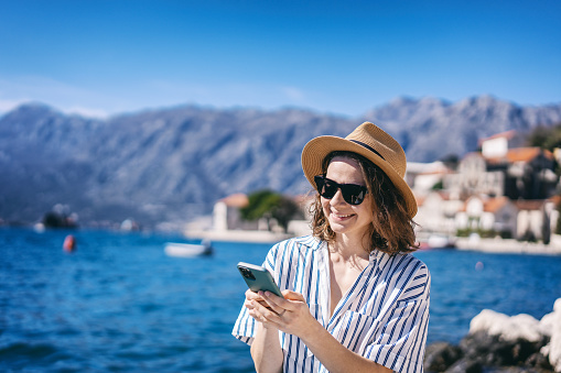 Young smiling woman in a hat taking a selfie using a smartphone on the shore of a mediterranean resort
