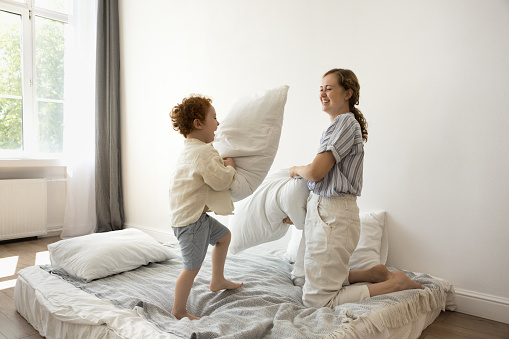 Joyful mom and toddler boy excited with pillow fighting on bed, having fun, laughing, shouting. Happy young mother and cute energetic kid son playing active games at cozy home