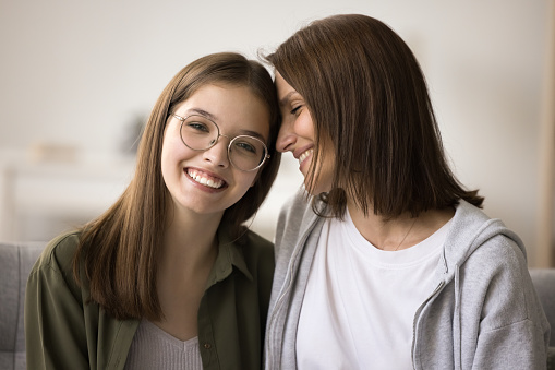 Cheerful teenage kid girl in glasses and happy mother sitting close on home sofa, hogging with love, affection, enjoying bonding, family relations, friendship, looking at camera with toothy smile