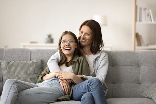 Happy mom and teenager daughter home casual portrait. Mother and teen girl resting on home sofa, hugging, talking with trust, having fun, laughing, enjoying bonding, friendship, relationships