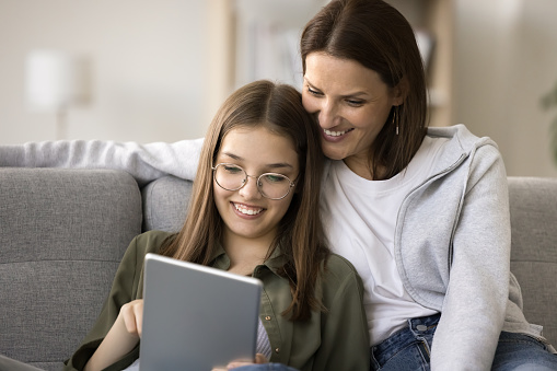 Cheerful pretty young mom and schoolgirl daughter in glasses using online service, educational application on tablet computer gadget, resting on home couch, shopping on Internet