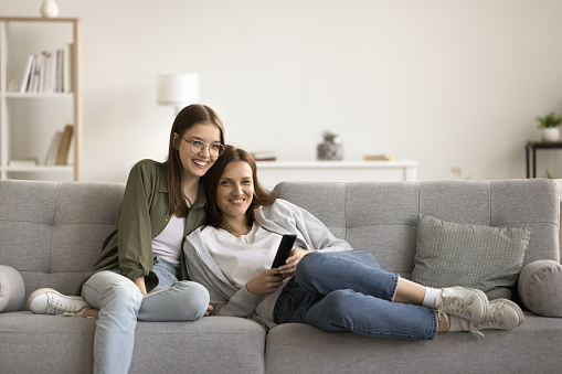 Cheerful mother and pretty teenager kid relaxing at cozy home, using mobile phone for Internet communication, resting on sofa with digital gadget, enjoying leisure, looking at camera