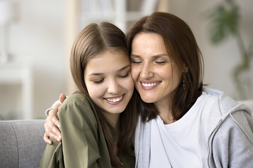 Loving young mother and happy teen daughter kid hugging with closed eyes and face touches, enjoying warm family moment, childhood, motherhood, expressing love, affection