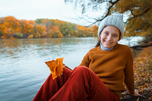 a young teenage girl posing in an autumn forest, sitting on a tree by the river bank, beautiful nature and bright yellow leaves