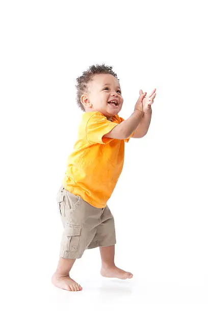 A full length image of a black toddler little boy wearing a bright orange shirt. He is standing and playfully clapping.