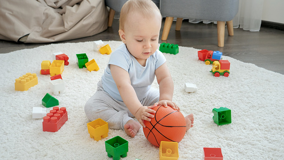 Little baby boy rolling and banging basketball ball on carpet at home. Concept of children development, sports, education and creativity at home.