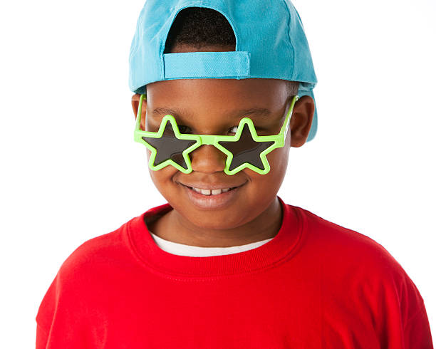 A closeup headshot of a smiling black little boy wearing a bright red shirt with a baseball cap and silly sunglasses.
