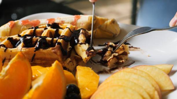 Enjoying Viennese Waffles Breakfast with Chocolate-Drizzled Fruits. Watch as the delightful breakfast spread of waffles, fruits, and chocolate syrup is sliced and savored with a knife and fork