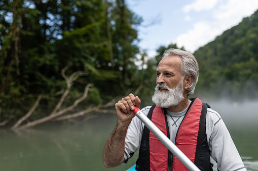 A gray-haired bearded man happily raises his oar in victory after paddling kilometers in a kayak and descends the green Kolpa river in Slovenia. Happy socializing on holidays and vacations.