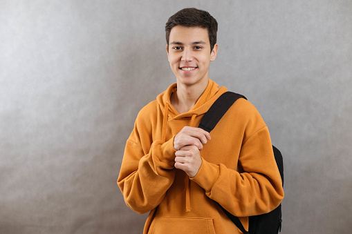 Portrait of a young student with a school bag. The teenager smiles and looks at the camera. A happy teenage boy on a grey background