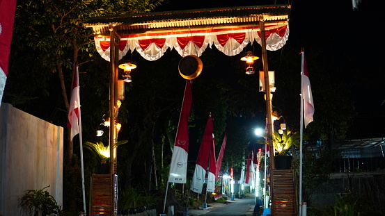 a bamboo gate with lanterns, some red and white decorations and banners on the side of the road to celebrate Indonesia's independence anniversary, photographed at night.