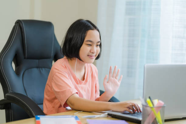 istock Asian kid saying hello before doing holiday homework online with laptop in front and using headphones to communicate with friends 1611910528