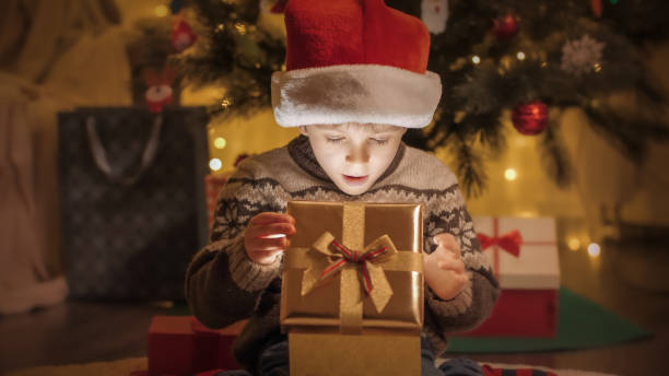 istock Happy smiling boy in sweater and Santa hat opens Christmas present box and looks inside 1611962006