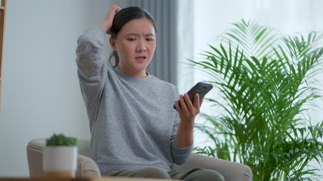 Asian woman feeling sadness reading or watching something at smartphone, sitting in living room at home.