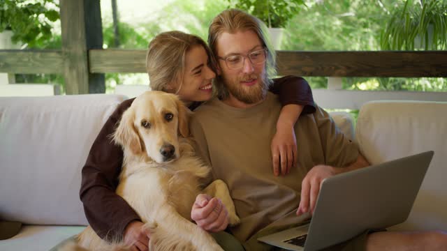 A happy family of a guy and a blonde girl, together with their dog, are considering goods in an online store on their laptop. Sitting on a sofa in a gazebo in nature
